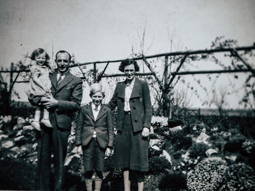 A vintage photograph features a family of four posing in a garden. The man on the left is holding a young child. A boy and a woman stand beside them. The background includes bushes and a wooden trellis with climbing plants.