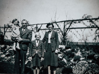 A vintage photograph features a family of four posing in a garden. The man on the left is holding a young child. A boy and a woman stand beside them. The background includes bushes and a wooden trellis with climbing plants.