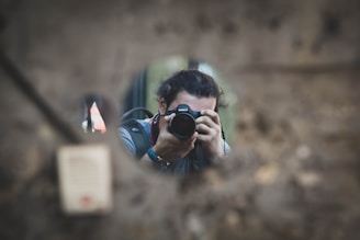 A person is taking a photograph through a small circular opening. The camera lens is pointed directly towards the viewer, and the scene is framed by blurred earthy textures, possibly a wall or natural material. The person's features are partially obscured by the camera they are holding.