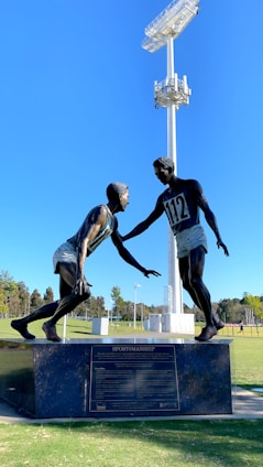 A sculpture depicting two athletes in mid-action is prominently displayed against a clear blue sky. One figure is reaching out to help the other, illustrating a moment of sportsmanship. The bronze statues stand on a black pedestal with an inscription, situated in an open green field with trees and sports facilities in the background. A tall floodlight tower looms overhead, adding to the outdoor sports setting.