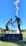 A sculpture depicting two athletes in mid-action is prominently displayed against a clear blue sky. One figure is reaching out to help the other, illustrating a moment of sportsmanship. The bronze statues stand on a black pedestal with an inscription, situated in an open green field with trees and sports facilities in the background. A tall floodlight tower looms overhead, adding to the outdoor sports setting.