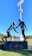 A sculpture depicting two athletes in mid-action is prominently displayed against a clear blue sky. One figure is reaching out to help the other, illustrating a moment of sportsmanship. The bronze statues stand on a black pedestal with an inscription, situated in an open green field with trees and sports facilities in the background. A tall floodlight tower looms overhead, adding to the outdoor sports setting.