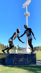 A sculpture depicting two athletes in mid-action is prominently displayed against a clear blue sky. One figure is reaching out to help the other, illustrating a moment of sportsmanship. The bronze statues stand on a black pedestal with an inscription, situated in an open green field with trees and sports facilities in the background. A tall floodlight tower looms overhead, adding to the outdoor sports setting.