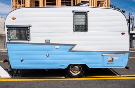A vintage travel trailer with a two-tone design, primarily in white and light blue, parked on a street. The side of the trailer features a window with blinds and a small emblem near the bottom. Behind the trailer, a wooden construction structure is visible.