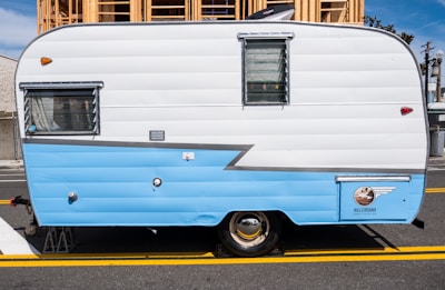 A vintage travel trailer with a two-tone design, primarily in white and light blue, parked on a street. The side of the trailer features a window with blinds and a small emblem near the bottom. Behind the trailer, a wooden construction structure is visible.