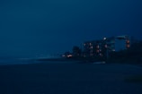 A nighttime view of a beach with an apartment building in the background. The building's windows emit a warm glow, contrasting with the deep blue sky. The ocean waves gently hit the shore, and a few small boats are visible on the sand nearby.