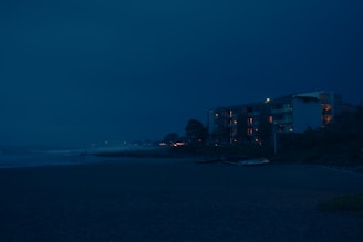 A nighttime view of a beach with an apartment building in the background. The building's windows emit a warm glow, contrasting with the deep blue sky. The ocean waves gently hit the shore, and a few small boats are visible on the sand nearby.