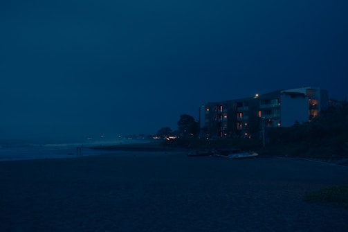 A nighttime view of a beach with an apartment building in the background. The building's windows emit a warm glow, contrasting with the deep blue sky. The ocean waves gently hit the shore, and a few small boats are visible on the sand nearby.