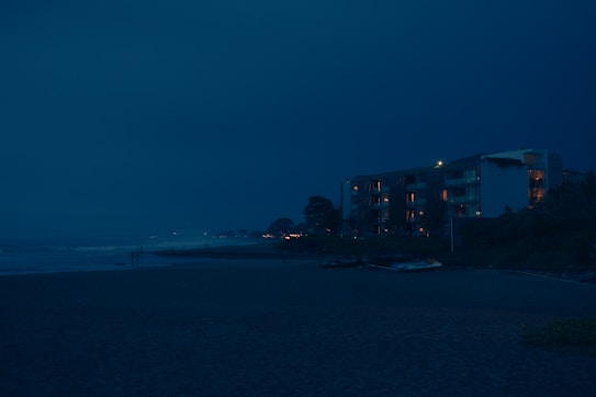 A nighttime view of a beach with an apartment building in the background. The building's windows emit a warm glow, contrasting with the deep blue sky. The ocean waves gently hit the shore, and a few small boats are visible on the sand nearby.