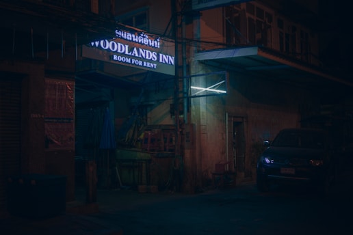 A dimly lit street corner featuring a sign for 'Woodlands Inn, Room for Rent'. The setting appears quiet and slightly worn, with a vehicle parked nearby. The building's exterior shows some signs of age and wear, while the surrounding area is mostly dark.