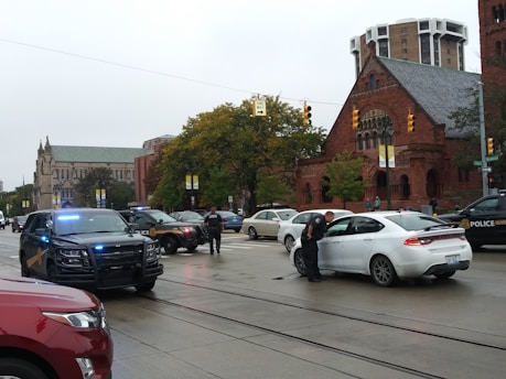 A city street is occupied by multiple police vehicles with blue lights flashing, surrounding a white car stopped at an intersection. Officers are standing near the vehicles, and a large, historic brick building is visible in the background along with several other architectural structures. Trees with autumn foliage line the street, contributing to an overcast and possibly rainy day.