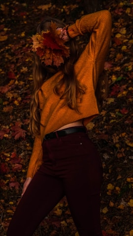 A person wearing a cozy orange sweater poses in a forested area covered with fallen autumn leaves. They are holding a cluster of vibrant orange and red leaves near their head, partially obscuring their face. The warm tones of the leaves and sweater create a harmonious blend with the autumnal setting.