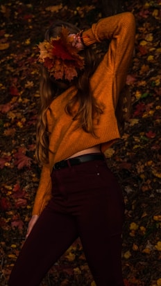 A person wearing a cozy orange sweater poses in a forested area covered with fallen autumn leaves. They are holding a cluster of vibrant orange and red leaves near their head, partially obscuring their face. The warm tones of the leaves and sweater create a harmonious blend with the autumnal setting.