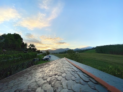A landscape featuring a foreground with a roof of overlapping stone tiles. In the background, a scenic view of fields stretches toward distant mountains under a partly cloudy sky illuminated by a setting or rising sun. Cars are parked along a driveway that runs parallel to a row of trees and bushes.