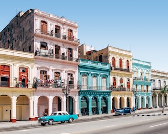 A row of colorful colonial-style buildings with balconies line a street. Each building features distinct pastel colors, including pink, blue, and yellow. Clothes hang on some balconies, and two vintage cars, one turquoise and the other blue, are parked on the street.
