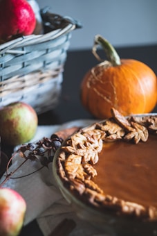 A rustic scene featuring a freshly baked pie with a detailed crust decorated with leaf designs. In the background, a wicker basket holds red apples and a small pumpkin is placed next to it. The setting conveys a cozy autumnal atmosphere.