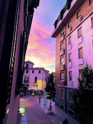 A vibrant urban scene featuring a narrow street flanked by tall buildings on either side. The sky is a striking mix of pink, purple, and blue hues, indicative of a sunset. In the background, people can be seen walking along the cobblestone street, with some sitting at an outdoor cafe. Streetlights cast a warm glow, adding to the evening atmosphere.