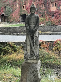 A stone statue of a knight in armor stands on a plinth amidst a garden setting, surrounded by green and red ivy-covered walls. The knight holds a sword that points downward, and the background features an old, brick facade with small windows.