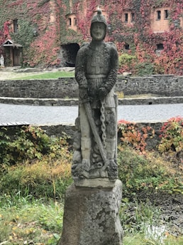 A stone statue of a knight in armor stands on a plinth amidst a garden setting, surrounded by green and red ivy-covered walls. The knight holds a sword that points downward, and the background features an old, brick facade with small windows.