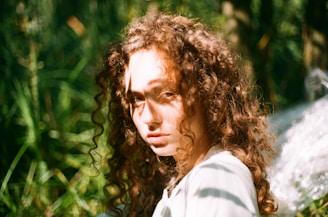 A person with curly hair is standing in a natural setting with sunlight filtering through leaves casting shadows on their face. The background is lush with green foliage, creating a serene and somewhat ethereal atmosphere.