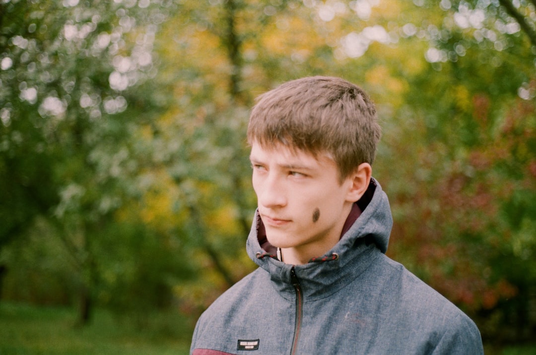 man wearing gray zip-up jacket standing while glancing his right side surrounded with tall and green trees