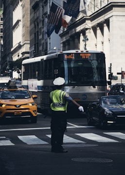 A police officer in a high-visibility vest directs traffic at a busy urban intersection. A bus is approaching in the background with a display sign reading 'BxM11 MIDTOWN 23 ST'. On the left, a yellow taxi is visible, and on the right, a dark-colored car can be seen. Several flags are hanging on a nearby building, and the architecture suggests a cityscape.