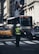 A police officer in a high-visibility vest directs traffic at a busy urban intersection. A bus is approaching in the background with a display sign reading 'BxM11 MIDTOWN 23 ST'. On the left, a yellow taxi is visible, and on the right, a dark-colored car can be seen. Several flags are hanging on a nearby building, and the architecture suggests a cityscape.