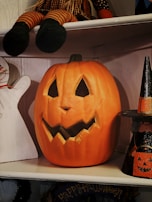 A carved pumpkin with a jagged smile and triangular eyes is sitting on a wooden shelf. Above it, a doll with striped legs is partially visible. On the right, a small witch decoration with a pointed hat is positioned. A ghost silhouette is partially visible on the left. The setup has a Halloween theme.