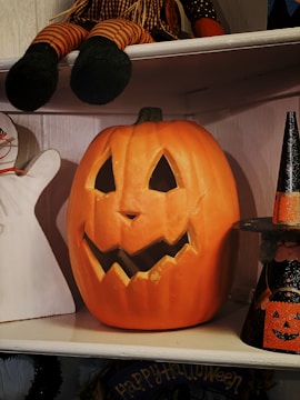 A carved pumpkin with a jagged smile and triangular eyes is sitting on a wooden shelf. Above it, a doll with striped legs is partially visible. On the right, a small witch decoration with a pointed hat is positioned. A ghost silhouette is partially visible on the left. The setup has a Halloween theme.