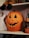 A carved pumpkin with a jagged smile and triangular eyes is sitting on a wooden shelf. Above it, a doll with striped legs is partially visible. On the right, a small witch decoration with a pointed hat is positioned. A ghost silhouette is partially visible on the left. The setup has a Halloween theme.