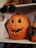 A carved pumpkin with a jagged smile and triangular eyes is sitting on a wooden shelf. Above it, a doll with striped legs is partially visible. On the right, a small witch decoration with a pointed hat is positioned. A ghost silhouette is partially visible on the left. The setup has a Halloween theme.