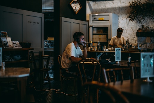 A dimly lit cafe interior with a warm, cozy atmosphere. A man sits at a table with a laptop and headphones, appearing focused and deep in thought. In the background, another person is working behind the counter, surrounded by various kitchen tools and shelves.
