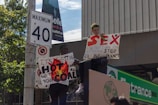 Two individuals are holding protest signs on a street corner. One sign reads 'SEX. Now that I have your attention, stop killing the planet,' and the other says 'It's getting hot in here so take off all your coal.' A speed limit sign and greenery are visible in the background.