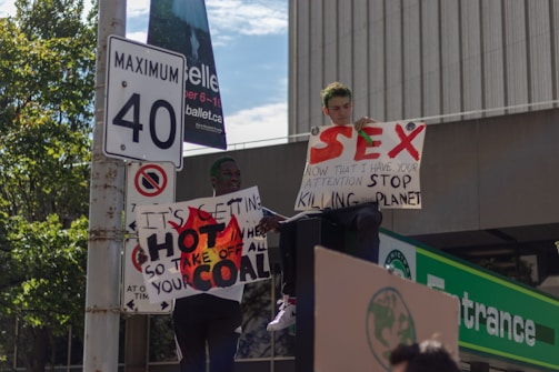 Two individuals are holding protest signs on a street corner. One sign reads 'SEX. Now that I have your attention, stop killing the planet,' and the other says 'It's getting hot in here so take off all your coal.' A speed limit sign and greenery are visible in the background.