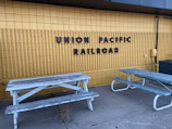 A yellow brick wall features bold, black lettering that spells 'Union Pacific Railroad.' In front of the wall, two weathered wooden picnic tables with peeling paint are situated on a cracked concrete surface. An air conditioning unit and some exposed cables are visible on the right side.