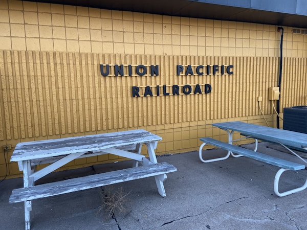 A yellow brick wall features bold, black lettering that spells 'Union Pacific Railroad.' In front of the wall, two weathered wooden picnic tables with peeling paint are situated on a cracked concrete surface. An air conditioning unit and some exposed cables are visible on the right side.