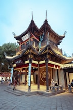A traditional Chinese building with intricate wooden architecture and curved tiled roofs. People are sitting and walking around, adding a lively atmosphere to the historic structure. Red and gold banners hang from the building, and there are Chinese characters visible on plaques.