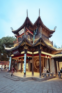 A traditional Chinese building with intricate wooden architecture and curved tiled roofs. People are sitting and walking around, adding a lively atmosphere to the historic structure. Red and gold banners hang from the building, and there are Chinese characters visible on plaques.