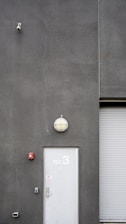 A utilitarian gray building exterior featuring a white door marked with 'TEC 3' and a dome-shaped light fixture above. A surveillance camera is mounted high on the wall, alongside a small, red fire alarm. A corrugated metal roller door is partially visible.
