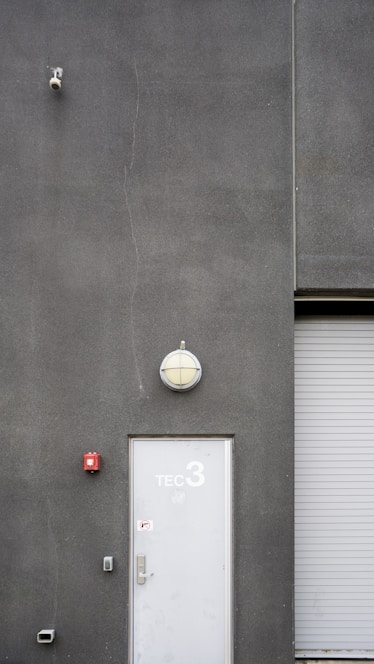 A utilitarian gray building exterior featuring a white door marked with 'TEC 3' and a dome-shaped light fixture above. A surveillance camera is mounted high on the wall, alongside a small, red fire alarm. A corrugated metal roller door is partially visible.