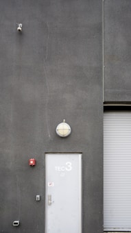 A utilitarian gray building exterior featuring a white door marked with 'TEC 3' and a dome-shaped light fixture above. A surveillance camera is mounted high on the wall, alongside a small, red fire alarm. A corrugated metal roller door is partially visible.