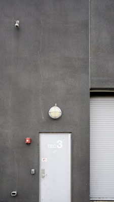 A utilitarian gray building exterior featuring a white door marked with 'TEC 3' and a dome-shaped light fixture above. A surveillance camera is mounted high on the wall, alongside a small, red fire alarm. A corrugated metal roller door is partially visible.