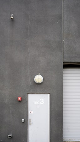 A utilitarian gray building exterior featuring a white door marked with 'TEC 3' and a dome-shaped light fixture above. A surveillance camera is mounted high on the wall, alongside a small, red fire alarm. A corrugated metal roller door is partially visible.