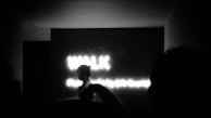A dimly lit scene with a silhouette of a person against a backdrop of illuminated text reading 'WALK'. The setting appears to be indoors, possibly in a corridor or tunnel, creating a mysterious atmosphere.