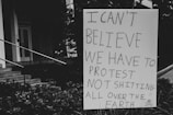 A sign with handwritten text expressing disbelief about needing to protest environmental issues is positioned outdoors near steps and foliage. The background includes part of a building facade and stairs.