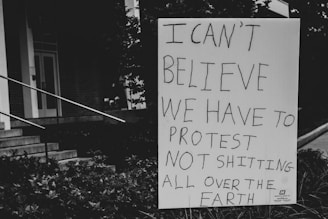 A sign with handwritten text expressing disbelief about needing to protest environmental issues is positioned outdoors near steps and foliage. The background includes part of a building facade and stairs.