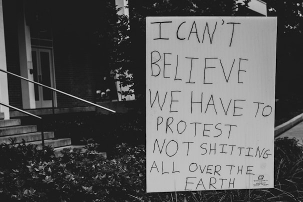 A sign with handwritten text expressing disbelief about needing to protest environmental issues is positioned outdoors near steps and foliage. The background includes part of a building facade and stairs.
