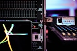 A close-up view of audio equipment, including an equalizer and a mixer, with a laptop displaying colorful waves on its screen. The scene is set in a dimly lit environment, emphasizing the glow of electronic displays and controls.
