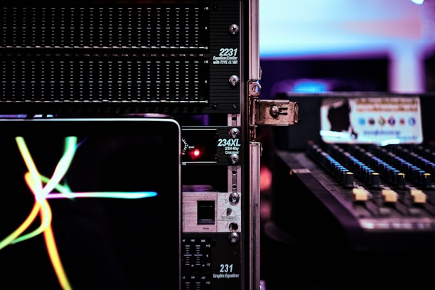 A close-up view of audio equipment, including an equalizer and a mixer, with a laptop displaying colorful waves on its screen. The scene is set in a dimly lit environment, emphasizing the glow of electronic displays and controls.