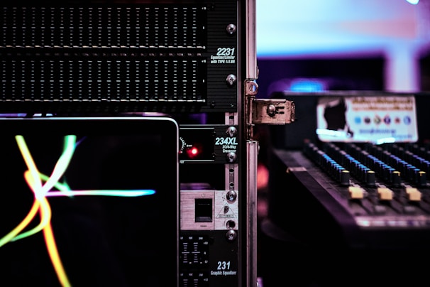 A close-up view of audio equipment, including an equalizer and a mixer, with a laptop displaying colorful waves on its screen. The scene is set in a dimly lit environment, emphasizing the glow of electronic displays and controls.